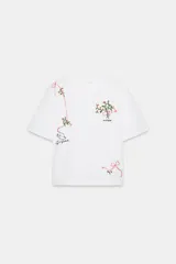 Camiseta blanca de manga corta y cuello redondo, con bordados de flores y lazos en tonos rosados y verdes. Incluye un bordado de un pájaro y texto en negro.