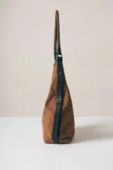 Cartera tipo hobo de gamuza color marrón, con correa trenzada de cuero negro y detalles laterales en cuero negro.