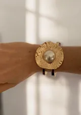 Brazalete rígido de diseño abierto, con una pieza central circular de textura martillada en tono dorado y una perla central.