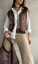 Chaleco de gamuza color marrón chocolate, con diseño sin mangas y escote en V. Cuenta con bordados en hilo claro y pequeños detalles de tachas en la parte frontal.