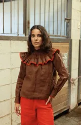 Camisa de lino color marrón, con cuello alto fruncido y detalle de volados en el pecho con borde en color rojo. Presenta mangas largas con puños abotonados.