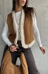 Chaleco de gamuza color camel con interior de corderito sintético, diseño sin mangas, borde frontal con ribete de corderito y detalle de tachas metálicas decorativas.