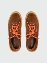 Zapatillas Moncler modelo Vera Leisure, confeccionadas en gamuza marrón con detalles en cuero tono sobre tono. Presentan cordones en color naranja vibrante y suela de goma texturizada en color caramelo.