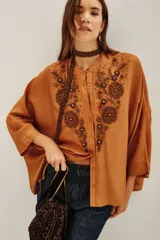 Camisa de viscosa color tostado, con corte holgado, mangas amplias y cuello mao. Presenta un elaborado bordado frontal con cuentas y pedrería en tonos oscuros.