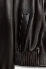 Cazadora tipo bomber de piel 100%, color marrón oscuro, con cuello subido y cierre frontal con cremallera metálica. Cuenta con bolsillos delanteros, forro interior y puños y bajo elásticos acanalados.
