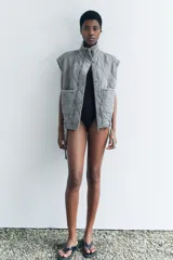 Chaleco gris acolchado con cuello alto, cierre frontal con cremallera metálica, bolsillos de plastrón en delantero y bajo ajustable con cordones.