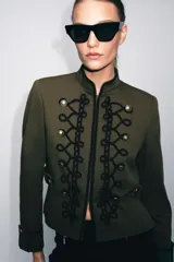 Blazer de estilo militar confeccionado en algodón, con cuello subido y manga larga. Presenta un diseño estructurado con detalle de alamares negros a contraste en el frente, botones metálicos decorativos y trabillas laterales. Cuenta con cierre frontal oculto y forro interior.