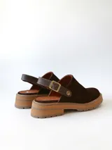 Zueco de cuero gamuzado color chocolate, con diseño de punta cerrada y tira ajustable con hebilla metálica en el talón. Presenta una suela de plataforma dentada en tono marrón claro.