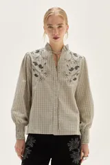 Blusa de algodón con estampado de rayas verticales en tonos crudo y negro. Presenta cuello mao con lazo para atar, bordados florales en negro y beige en la zona del pecho y hombros, y mangas largas con puños fruncidos elásticos.