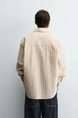 Camisa de algodón de corte relajado, color beige con finas rayas verticales en tonos gris y amarillo. Presenta cuello solapa, manga larga con puños abotonados y bajo con aberturas laterales.