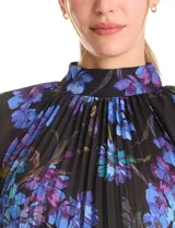 Blusa de gasa plisada con estampado floral en tonos azules y violetas sobre fondo negro. Presenta cuello alto, mangas largas con puños abotonados y un diseño fluido con textura plisada.
