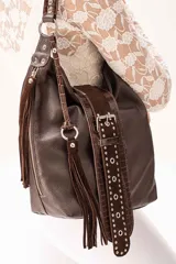 Cartera de hombro color chocolate, confeccionada en una combinación de cuero vacuno graneado, cuero grabado tipo croco y gamuza. Presenta un diseño con flecos laterales, una tira central decorativa con tachas metálicas y hebilla funcional, cierre principal metálico y correa larga regulable y desmontable.
