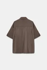 Camisa de corte fluido con cuello solapa y manga corta con dobladillo. Presenta un cierre frontal con botones ocultos bajo una tapeta y un diseño con bajo ligeramente más largo en la parte posterior.