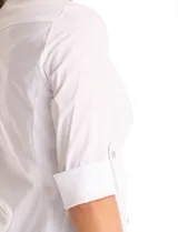 Camisa blanca de algodón con cuello solapa, escote en V y cierre frontal con botones decorativos de strass. Presenta pinzas de entalle para un ajuste definido, mangas tres cuartos con terminación rebatible y ruedo redondeado.