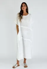 Vestido midi de lino color blanco, con corte holgado, cuello redondo y mangas cortas. Presenta detalles de costuras horizontales con terminaciones desflecadas en el frente y el ruedo.