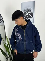 Campera de jean azul oscuro con capucha, cierre frontal y bolsillos tipo canguro. Presenta un bordado floral de flores de cerezo en el pecho derecho.