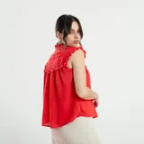 Camisa sin mangas color beige, con cuello alto con volados, detalles de encaje calado en los hombros y parte superior del pecho, y cierre frontal con botones.