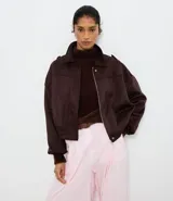 Campera bomber de gamuza sintética color marrón oscuro, con cuello clásico, cierre frontal, hombros caídos y puños ajustados. Presenta un diseño holgado con textura aterciopelada.