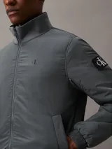 Campera bomber gris de nailon reciclado, con cuello alto, cierre de cremallera, bolsillos laterales y puños y bajo elásticos.