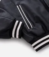 Campera bomber de cuero sintético con diseño bicolor, cuerpo negro y mangas blancas. Presenta cuello, puños y cintura con tejido elástico a rayas, cierre frontal con botones a presión y bordado con la palabra Authentic en el pecho.