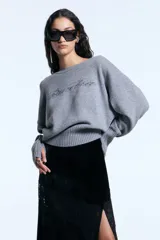 Sweater de tejido de punto gris con calce holgado, cuello redondo y mangas largas con puños que incluyen orificio para el pulgar. Presenta un diseño de letras bordadas con aplicación de strass en el frente y terminaciones en ribb ancho.