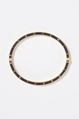 Pulsera rígida de metal dorado con detalles de esmalte negro en el borde y letras grabadas a lo largo de la pieza. Cuenta con cierre de bisagra.