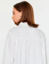 Camisa blanca de manga larga con cuello clásico y cierre frontal de botones. Presenta detalles bordados florales tono sobre tono en la zona de los hombros y el pecho.
