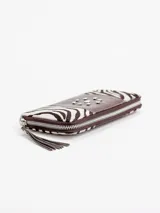Billetera rectangular de cuero con cierre perimetral, diseño combinando laterales de pelo con estampado animal print tipo cebra y franja central roja con tachas metálicas dispuestas en forma circular. Cuenta con tirador de cierre en flecos de cuero rojo.