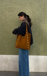 Cartera tipo tote de símil gamuza color camel con diseño amplio, correas de hombro integradas y detalles de tachas metálicas en los laterales.