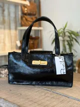 Cartera tipo tote bag de diseño rectangular, confeccionada en cuero negro con acabado brillante y efecto arrugado. Presenta doble asa de mano, cierre frontal con solapa y herraje metálico dorado tipo torniquete.