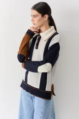 Buzo de punto con cuello polo y cierre frontal de tres botones. Presenta un diseño a rayas verticales en tonos beige y marrón, con mangas en bloques de color rojo y detalles en rosa en los puños y el ruedo.