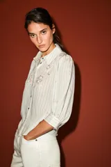 Camisa blanca a rayas sutiles, de cuello solapa y manga abullonada por debajo del codo. Presenta delicados bordados florales perforados en el frente y cierre frontal con botones.