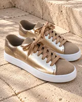 Zapatillas urbanas de cuero metalizado dorado y gamuza beige, con cordones anchos al tono y suela blanca.