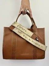 Cartera marrón de cuero genuino con doble asa corta y correa bandolera estampada con la frase "Chabas, soy real, no perfecta".