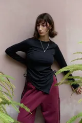 Top de manga larga color negro, confeccionado en jersey elástico. Presenta un diseño asimétrico con jaretas laterales regulables que permiten ajustar el largo y el volumen, y aberturas para los pulgares en los puños.