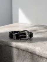 Cinto fino de color negro con textura tipo reptil y hebilla metálica plateada con diseño grabado estilo western.