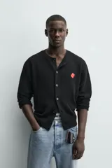 Cárdigan de punto negro, corte relajado, con cuello redondo, manga larga y cierre frontal de botones oscuros. Presenta un parche bordado en forma de dado rojo con corazones blancos en el lado izquierdo del pecho.