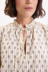 Camisa de manga larga color amarillo pálido con estampa boho a rayas verticales compuestas por pequeños motivos florales. Posee cuello alto con volados y cordones para atar en el frente, y mangas abullonadas con puños ajustados.