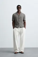 Camisa de punto de manga corta con cuello bowling y cierre frontal de botonadura. Presenta un patrón de rayas horizontales finas en blanco y negro, con ribetes negros en cuello, puños y bajo.