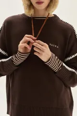 Sweater de tejido de punto color beige, con cuello redondo y detalles de bordado en zigzag en color azul oscuro a lo largo de las mangas y hombros. Presenta un pequeño bordado con texto en el pecho.