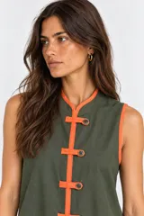 Vestido corto sin mangas color verde militar con ribetes y alamares en color naranja, cuello mao, cierre frontal con botones y volado en el ruedo.