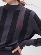 Sweater de punto con diseño de rayas verticales en tonos negro y gris oscuro. Presenta cuello alto tipo polera, mangas largas con puños ajustados y ruedo elástico. Incluye un pequeño bordado en color rojo en el pecho.