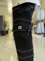 Pantalón cargo de jean negro con múltiples bolsillos utilitarios y costuras visibles en contraste de color blanco.