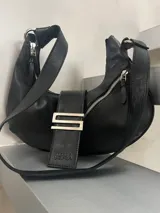 Cartera de cuero genuino color negro, con forma de media luna (hobo bag). Cuenta con asa regulable para llevar al hombro o cruzada, cierre principal de cremallera y broche imán. Presenta un herraje metálico distintivo en forma de "S" en la correa frontal.