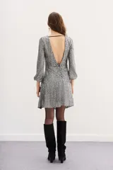 Vestido corto de fiesta con lentejuelas plateadas en patrón de espiga, mangas tres cuartos y cuello redondo. El diseño presenta un ajuste entallado con un cinturón ancho negro de gamuza con hebilla metálica dorada en forma de corazón.