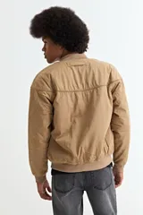 Campera tipo bomber color tostado, con cierre frontal, cuello, puños y ruedo de tejido elástico acanalado. Presenta costuras horizontales en la parte superior del pecho.