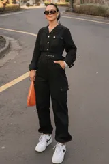 Mono largo de gabardina color negro, de manga larga, con cuello camisero, cierre frontal con botones metálicos, bolsillos con solapa en el pecho y estilo cargo con bolsillos laterales en las piernas. Incluye un cinturón ancho ajustable con hebilla forrada.