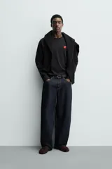 Camiseta de manga larga color negro, corte relajado (relaxed fit) con cuello redondo y un pequeño parche bordado en el pecho con un dado rojo y corazones blancos.