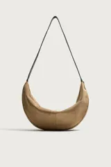 Bolso de hombro liso de diseño curvado y estilo hobo, confeccionado en material tipo gamuza color tostado, con una correa larga y fina en color negro. Cuenta con un compartimento principal con cierre de cremallera y un bolsillo interior con cremallera.