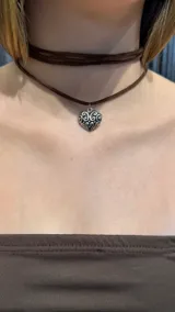 Gargantilla tipo choker de gamuza marrón con diseño de doble vuelta y dije de corazón metálico plateado con detalles labrados.